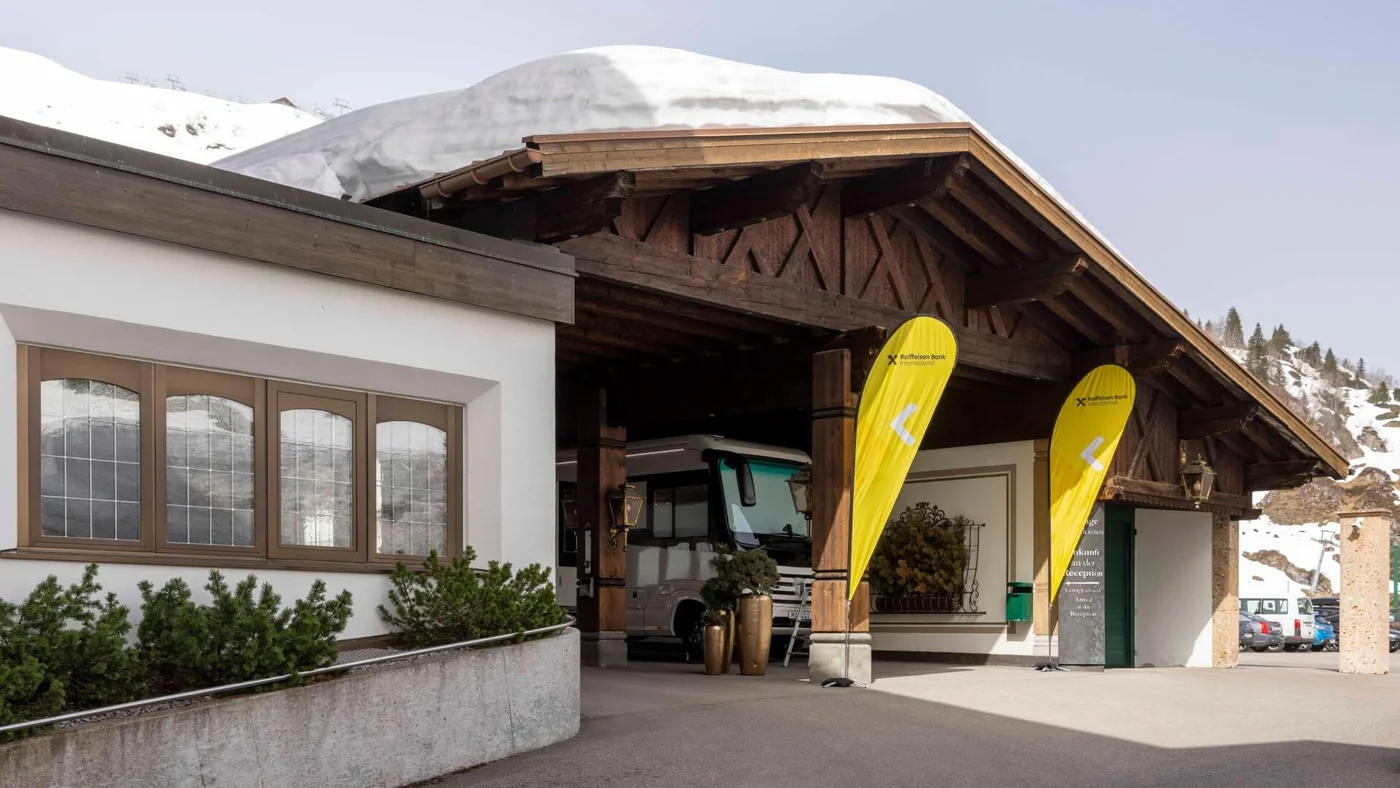 Der Eingang eines Berghotels mit traditioneller Holzarchitektur, Schnee auf dem Dach und gelben Fahnen mit Logo davor.