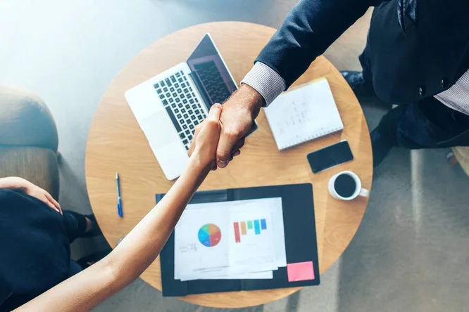 Zwei Fachleute schütteln sich an einem runden Bürotisch mit Laptop, Finanzdiagrammen, Notizbuch und Kaffeetasse die Hände und symbolisieren damit ein erfolgreiches Geschäftsabschlüsse oder eine erfolgreiche Partnerschaft.