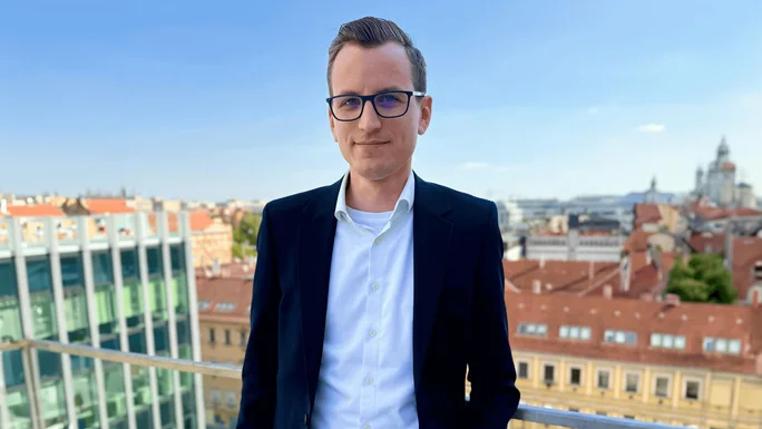 A person dressed in a dark suit and white shirt stands on a balcony overlooking a cityscape. The scene captures a clear blue sky and urban architecture.