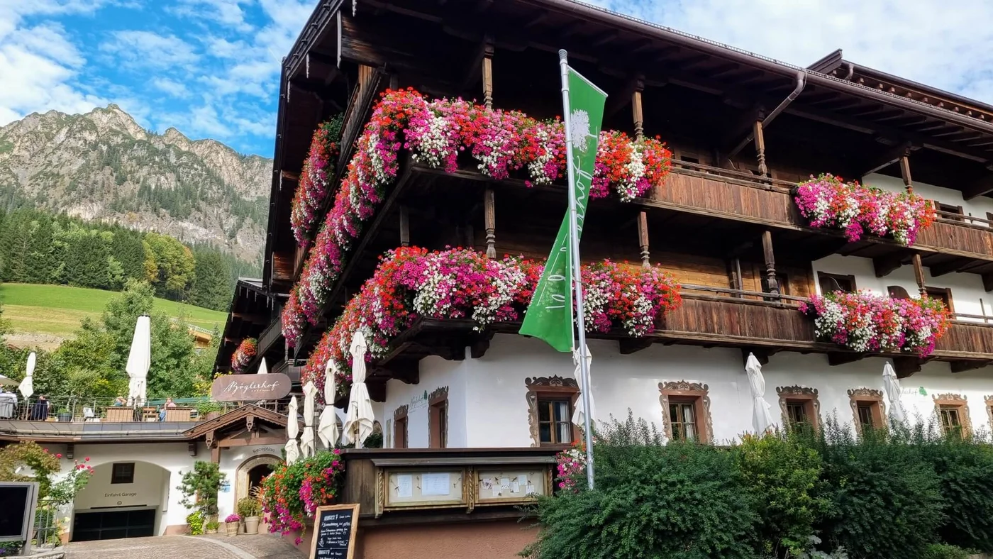 Ein alpines Holzgebäude mit bunten Blumen auf den Balkonen, vor der Kulisse einer Berglandschaft und einer grünen Landschaft.