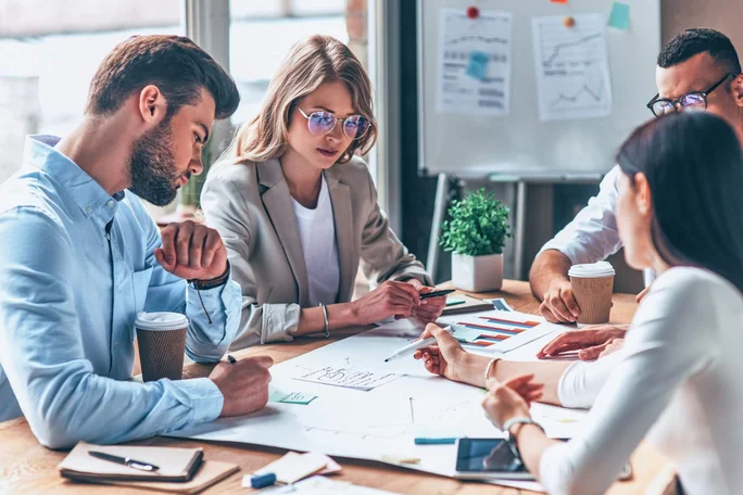 Eine Gruppe von Fachleuten diskutiert aktiv und entwickelt Strategien an einem mit Diagrammen und Grafiken bedeckten Tisch und fördert so Teamarbeit und Innovation in einer modernen Büroumgebung.