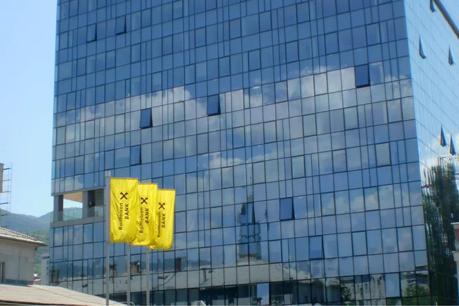 Moderne Glasfassade eines hohen Gebäudes mit deutlich sichtbaren Raiffeisen-Bank-Fahnen im Vordergrund. Die spiegelnde Oberfläche des Gebäudes zeigt einen klaren blauen Himmel mit Wolken.