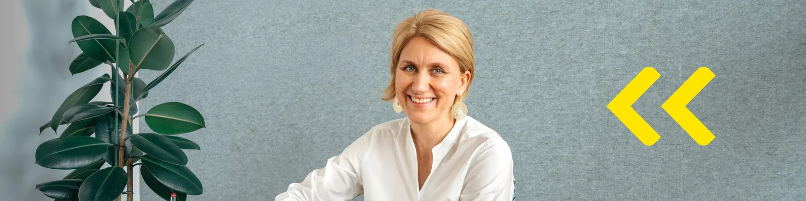 A professional woman with blonde hair sits in a modern office setting against a gray wall. Her face is blurred for privacy, and she wears a white shirt. A large green indoor plant is visible on the left side of the image. Two bright yellow chevron arrows appear on the right, adding a graphic design element and sense of direction.