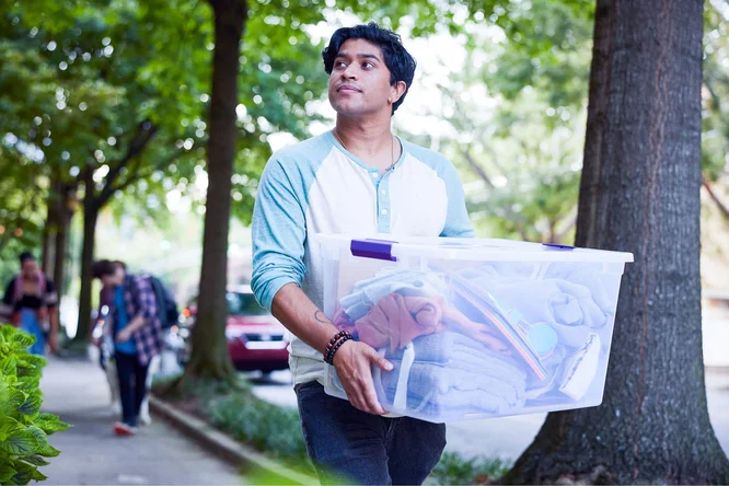 An individual carrying a large plastic container filled with household items, walking along a tree-lined street, symbolizing the process of moving or relocating.