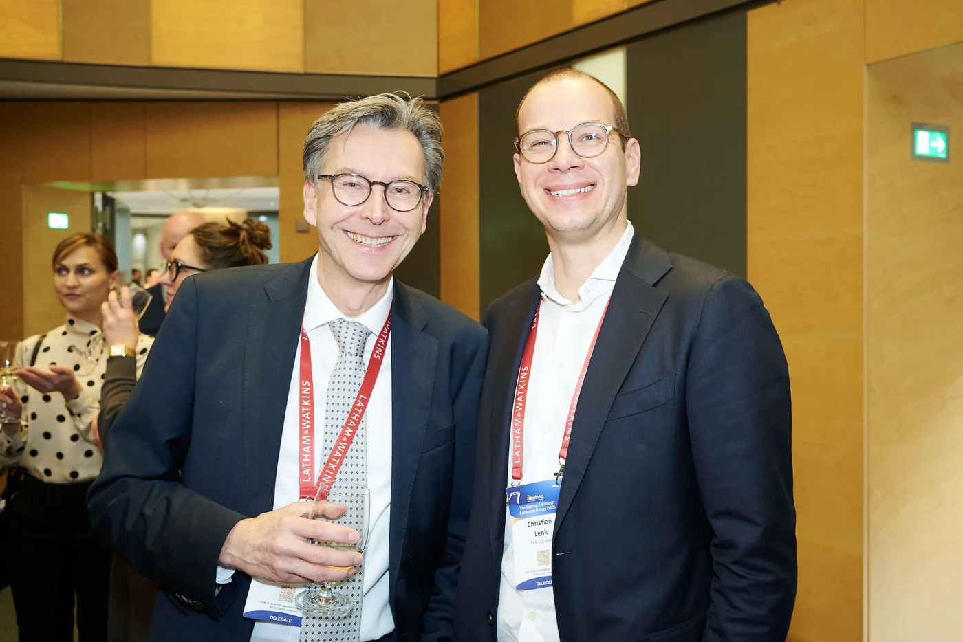 Zwei Männer in Businesskleidung mit Konferenz-Lanyards und Namensschildern stehen gemeinsam bei einer Netzwerkveranstaltung.