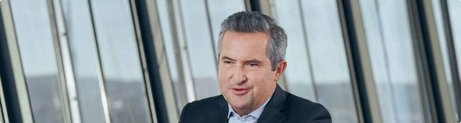 Rainer Schnabl sitzt in einem modernen Büro. Er trägt ein dunkles Sakko und ein helles Hemd, was auf ein formelles Geschäftsumfeld hindeutet. Große, vertikale Glasfenster im Hintergrund geben den Blick auf eine urbane oder industrielle Landschaft frei. Die Szene vermittelt Professionalität und eine moderne Arbeitsatmosphäre.