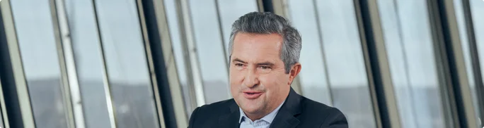 Rainer Schnabl is seated in a contemporary office environment. The person is dressed in a dark suit jacket and light shirt, suggesting a formal corporate setting. Large vertical glass windows in the background reveal an urban or industrial landscape. The overall scene conveys professionalism and a modern workplace atmosphere.