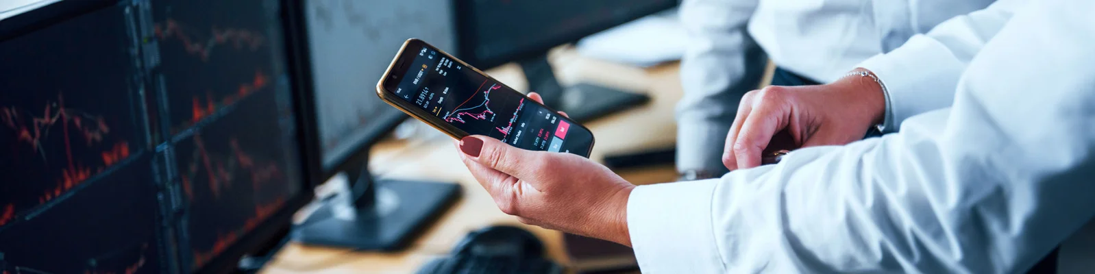 Ein Fachmann überprüft Finanzdaten auf einem Smartphone, während er von mehreren Computerbildschirmen umgeben ist, die Aktienmarktcharts anzeigen. Das Bild vermittelt einen Fokus auf Echtzeitanalyse und Entscheidungsfindung.