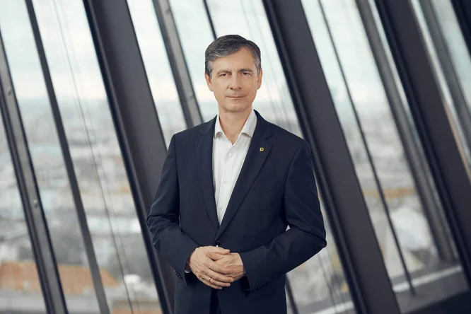Michael Alge steht selbstbewusst in einem dunklen Anzug in einem modernen Büro mit Glaswänden. Mit leicht gefalteten Händen blickt er in die Kamera und trägt ein weißes Hemd. Hohe Fenster im Hintergrund geben den Blick auf eine unscharfe Stadtkulisse frei. Sanftes, neutrales Licht und gedeckte Farben schaffen eine professionelle und ruhige Atmosphäre. Die Szene wirkt wie ein Porträt einer Führungskraft in einem Unternehmen.