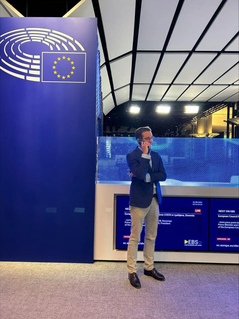 Paul Pasquali in business attire stands with arms crossed in a modern institutional interior. He is positioned beside a large blue wall featuring the European Union flag and a circular emblem. Behind him, illuminated digital screens display text and program information in a blue color scheme. The setting appears to be a contemporary media or broadcast area within a parliamentary or conference environment.