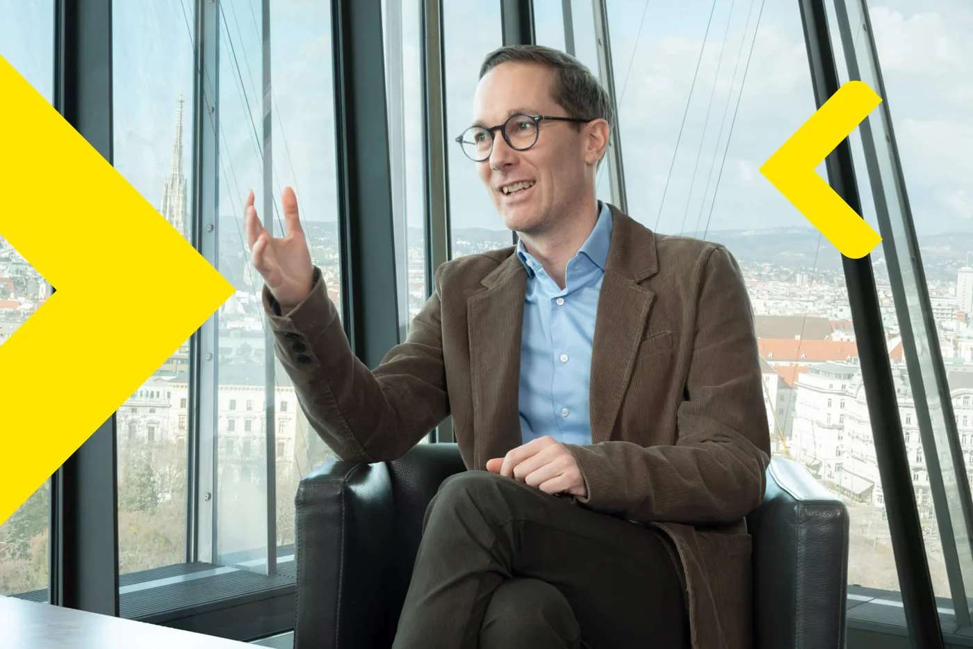 Michael Heinzl in a brown blazer and blue shirt sits in a contemporary office with large glass windows, gesturing as he speaks. The background reveals a cityscape with buildings and a partly cloudy sky. Bright yellow geometric shapes overlay the image, adding a dynamic visual element. The atmosphere is professional and modern, with natural daylight illuminating the scene.