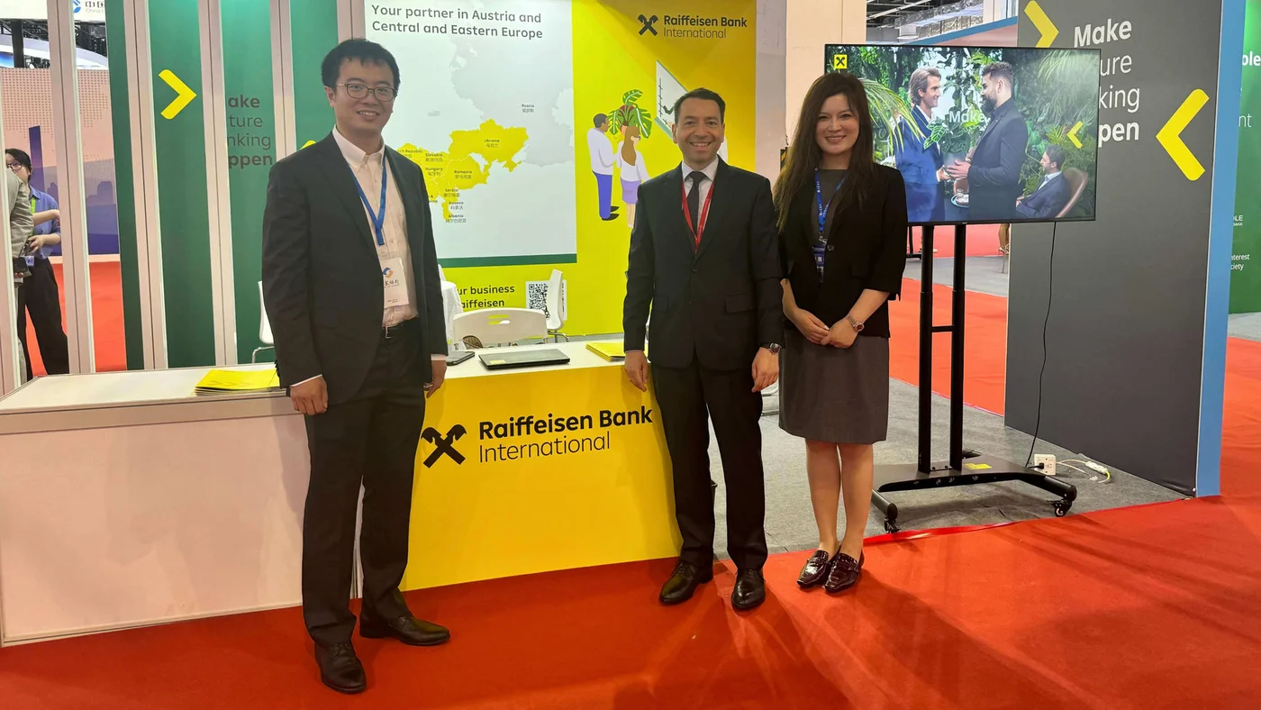 A trade show booth showcasing Raiffeisen Bank International with branding prominently displayed. The setting includes a bright yellow backdrop, informational panels, and a digital screen. Three individuals are standing near the booth, dressed in formal attire. The booth highlights the bank's presence in Austria and Central Europe.