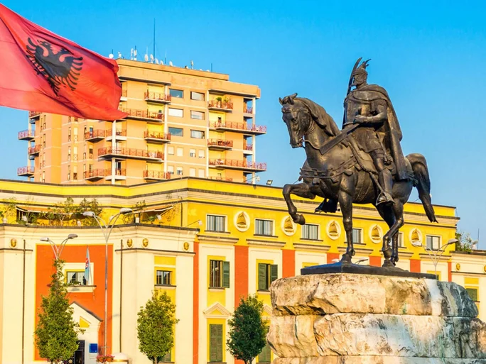 A prominent statue of Skanderbeg, the national hero of Albania, mounted on horseback in a public square. The Albanian flag waves nearby, adding a patriotic touch to the scene. The background features colorful architecture and a residential building under a clear blue sky.
