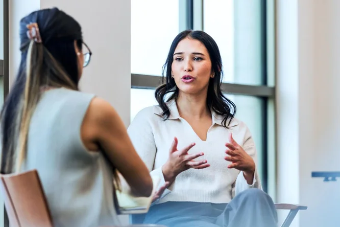 Zwei Frauen führen in einem modernen Büroumfeld ein berufliches Gespräch, bei dem Zusammenarbeit, Kommunikation und Teamwork im Vordergrund stehen.