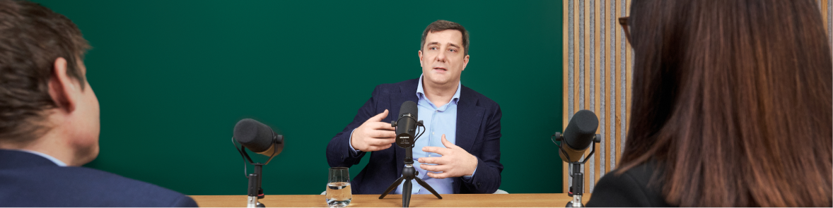 A person in a suit speaks into a microphone at a table, facing two other individuals. The setting appears to be a formal meeting or interview, with a green background and wooden paneling. Two microphones and a glass of water are visible on the table, suggesting a professional or academic environment. The overall mood is serious and focused.