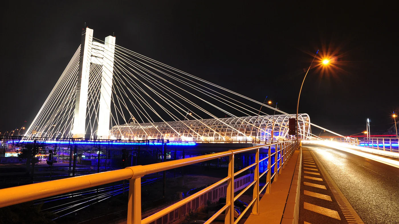 Eine markante Schrägseilbrücke erstrahlt im nächtlichen Himmel, helle Lichter und Spiegelungen schaffen eine faszinierende Szenerie. Auf der Fahrbahn sind Lichtspuren vorbeifahrender Fahrzeuge zu sehen.