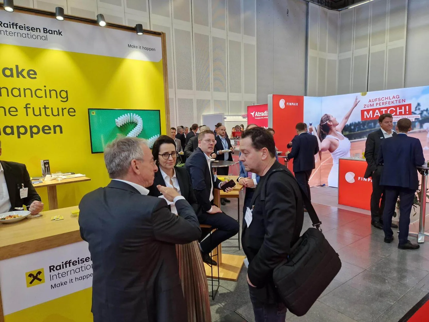 Messestand der Raiffeisen Bank International auf einer Fachmesse mit leuchtend gelbem Hintergrund und dem Slogan „Make financing the future happen“. Besucher:innen vernetzen sich und führen Gespräche – dies betont die dynamische und interaktive Atmosphäre der Veranstaltung.