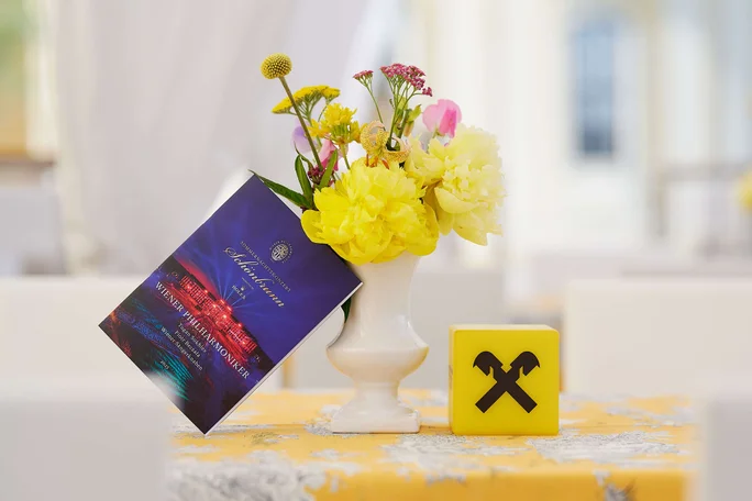 A white vase with colorful flowers, a brochure hanging on the vase, and a yellow block with black logo on a table covered with a yellow and white tablecloth.
