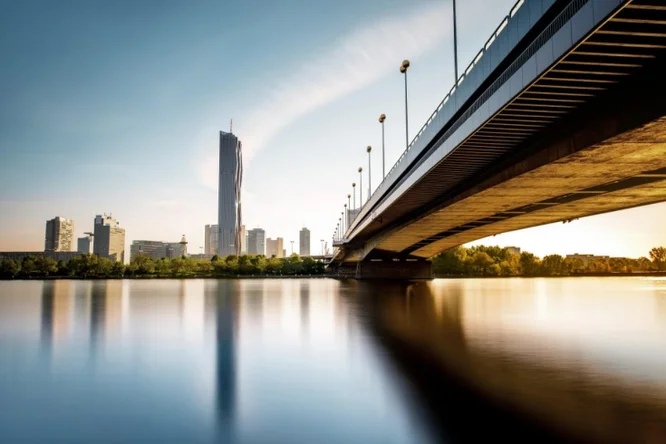 Eine moderne Stadtsilhouette mit hohen Gebäuden und einer Brücke, die über einen ruhigen Fluss bei Sonnenuntergang führt.
