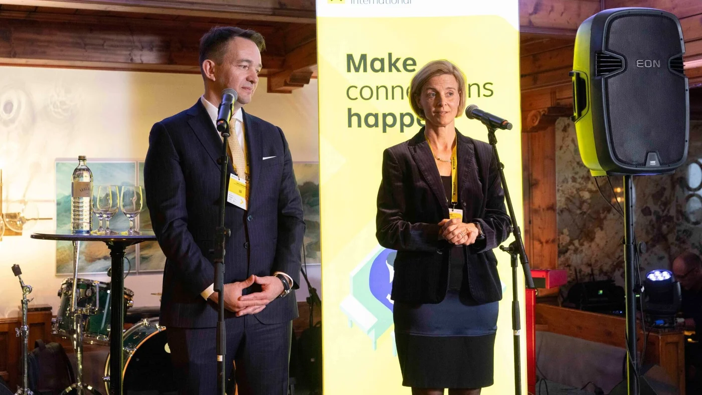 Ein Mann und eine Frau stehen bei einer Konferenz an Mikrofonen, vor einem gelben Banner mit dem Text "make it happen" und Branding-Elementen. Der Hintergrund zeigt ein Holzinterieur mit Musikinstrumenten und Beleuchtung.
