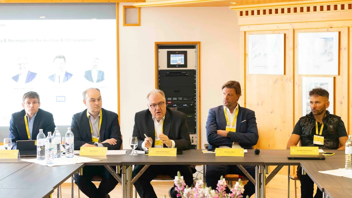 Eine Gruppe von fünf Berufstätigen in Anzügen sitzt bei einer Konferenz an einem Tisch, mit einem großen Bildschirm und Branding im Hintergrund, und führt eine Diskussion oder Präsentation.