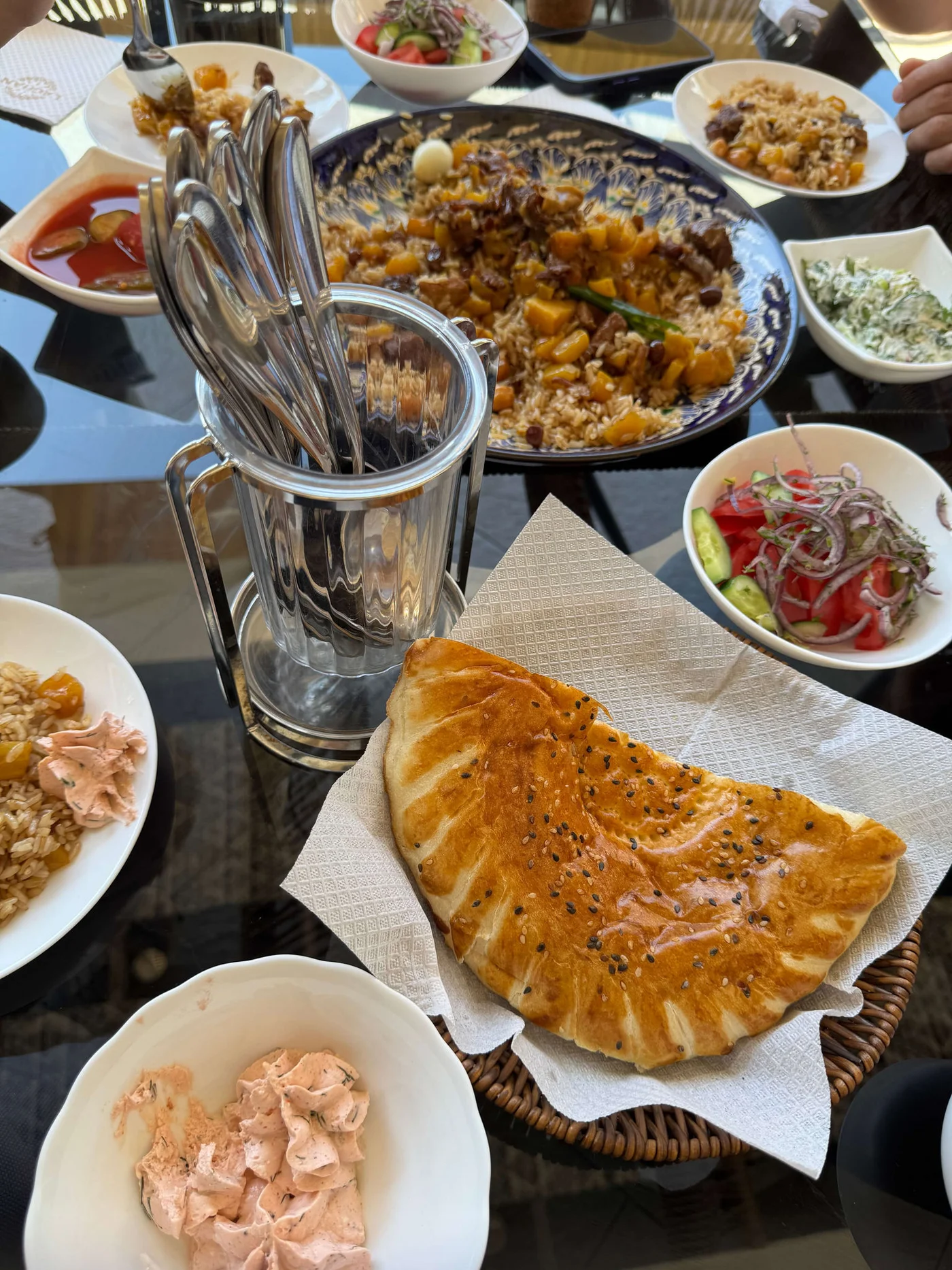 A variety of Middle Eastern dishes are arranged on a glass table, centered around a large plate of rice and meat. A golden, sesame-topped flatbread rests on paper towels in a wicker basket in the foreground. Small bowls of salads, spreads, and grains surround a tall glass filled with cutlery. The scene suggests a shared meal in a bright, casual dining setting.