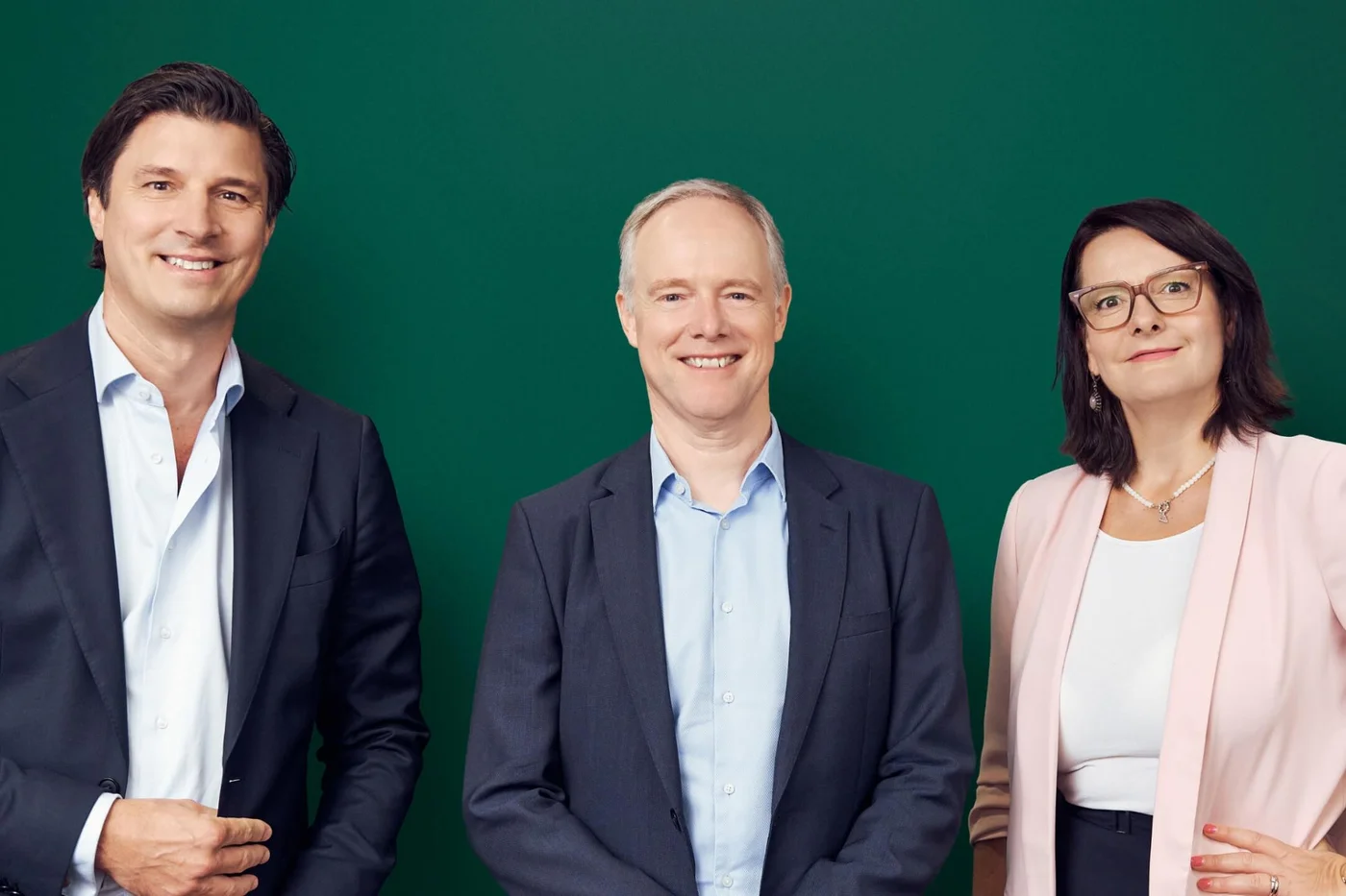 Three professionally dressed adults stand side by side in front of a solid green background. Two men in dark suits and a woman in a light blazer pose confidently, facing the camera. Their expressions are friendly and approachable, suggesting a corporate or executive setting. The clean backdrop and formal attire emphasize a modern, professional image suitable for business or leadership themes.