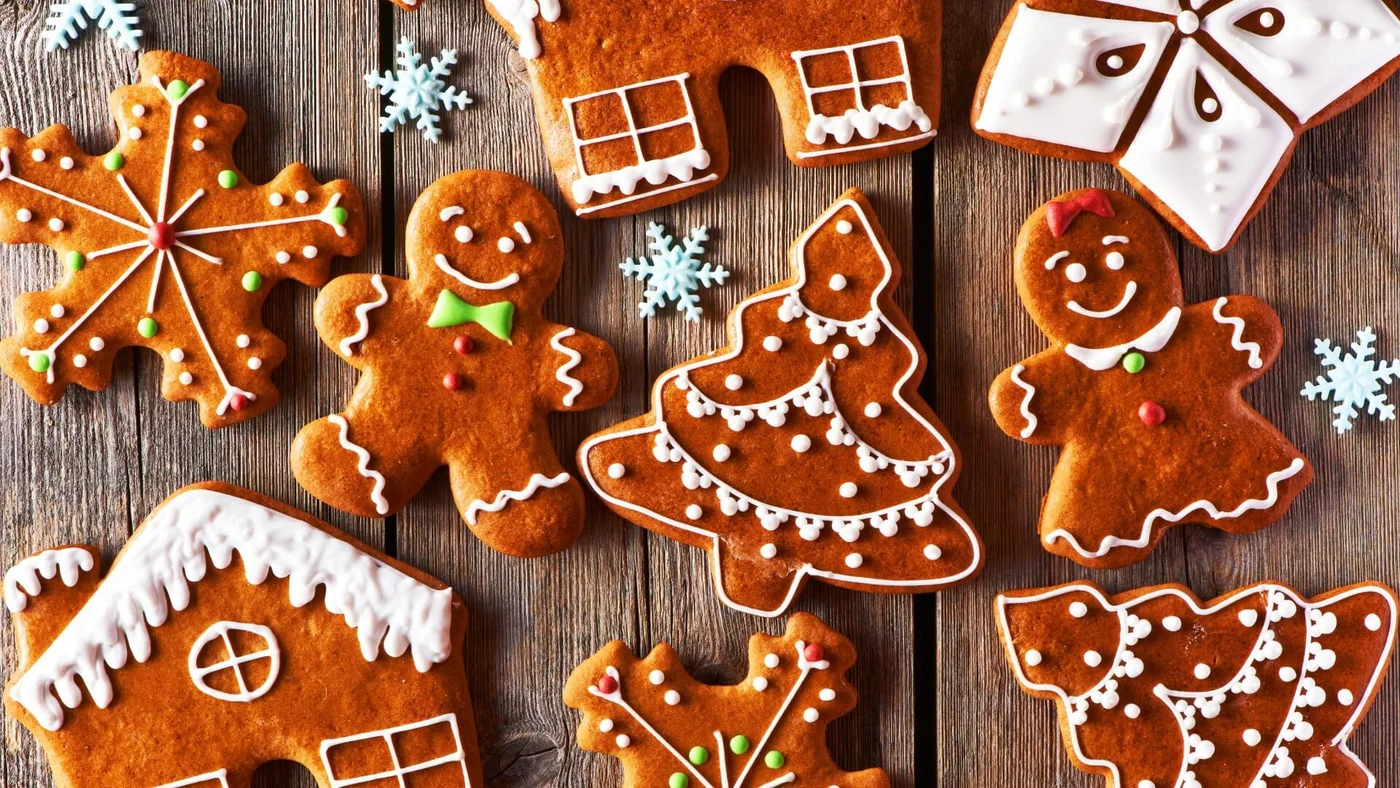 Verschiedene Lebkuchen mit weißen, grünen und roten Zuckergussdekorationen, in Formen von Schneeflocken, Lebkuchenmännern, Weihnachtsbäumen und Häusern, auf einer Holzoberfläche.