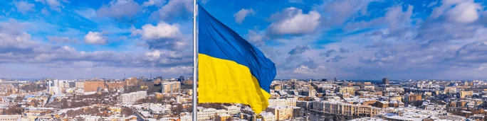 The Ukrainian flag waves prominently against a backdrop of a cityscape under a vibrant blue sky with scattered clouds.