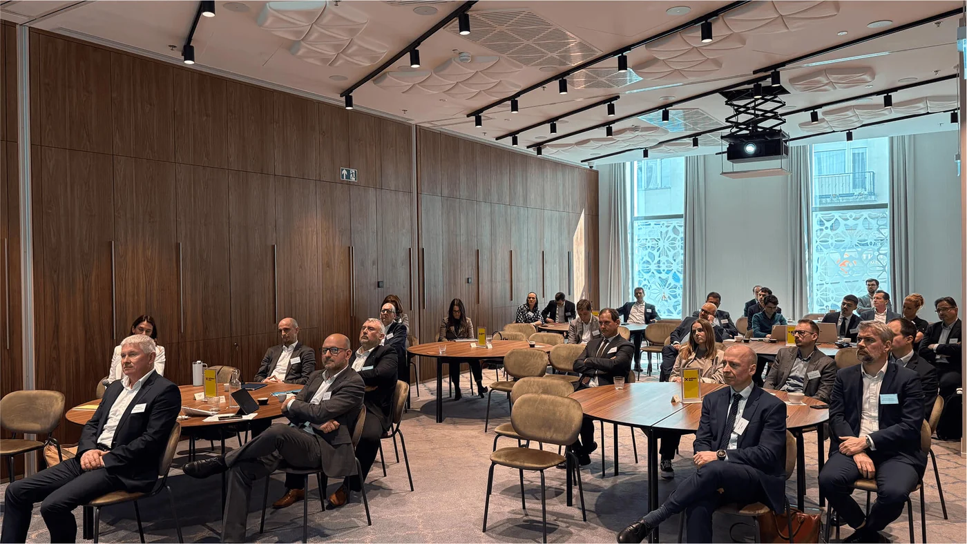 Attendees seated at round tables during a business conference in a contemporary room with wooden walls and large windows. The setting includes a projector for presentations.