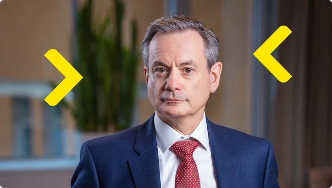 Laszlo Havas professionally dressed, wearing a navy blue suit, white shirt, and red tie is seated indoors. The background features a blurred office setting with a plant and large windows. Yellow arrow graphics are overlaid on the image, adding a modern design element.