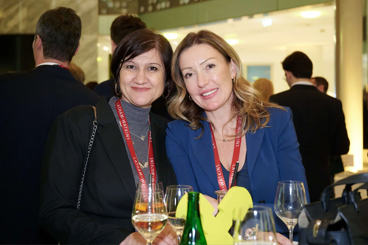 Zwei elegant gekleidete Frauen bei einer geschäftlichen Netzwerkveranstaltung, gemeinsam mit Weingläsern und Event-Lanyards.