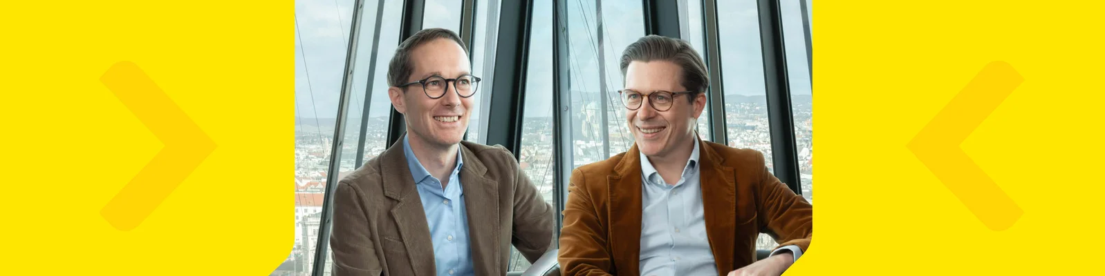 Björn Zaksek and Michael Heinzl are seated indoors near large glass windows with a cityscape visible in the background. Both are wearing brown jackets and light blue shirts, suggesting a professional or business casual setting. The scene is brightly lit, with a yellow graphic border and arrows on each side. The overall mood is modern and professional, with a focus on conversation or interview.