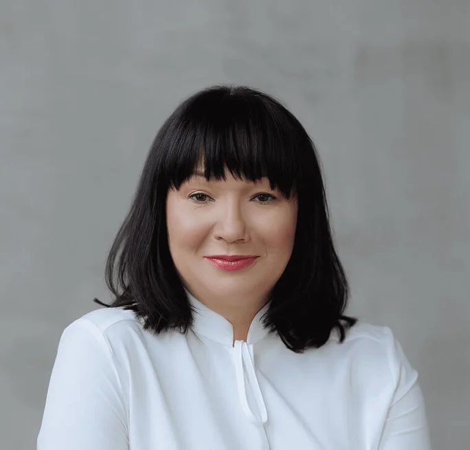 A person with shoulder-length black hair, wearing a white blouse, set against a neutral gray background. The image conveys a formal and professional appearance.