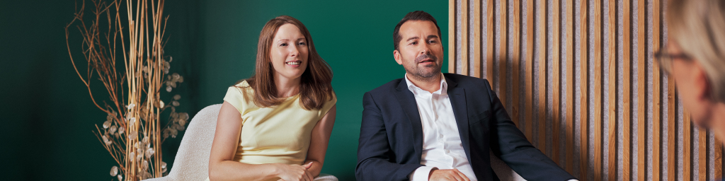 Two individuals are seated in a modern office environment, engaging in a professional discussion. The setting features a green wall, wooden paneling, and minimalist decor with dried plants. The individuals are dressed in formal and semi-formal attire, suggesting a business or consultation scenario.