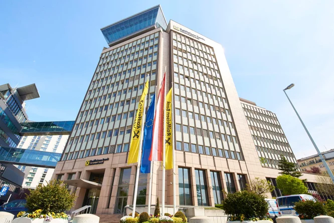 Das Gebäude der Raiffeisen Bank International mit moderner Architektur, großen Glasfenstern und Fahnen davor, unter klarem, blauem Himmel.