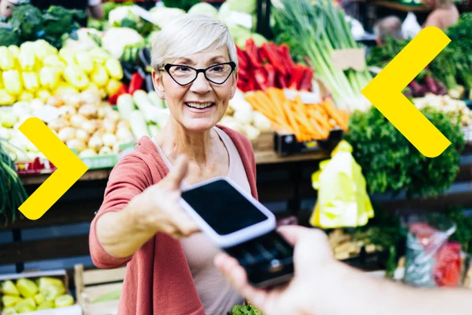 Smiling older woman using SoftPOS to accept a contactless payment at a vegetable market.