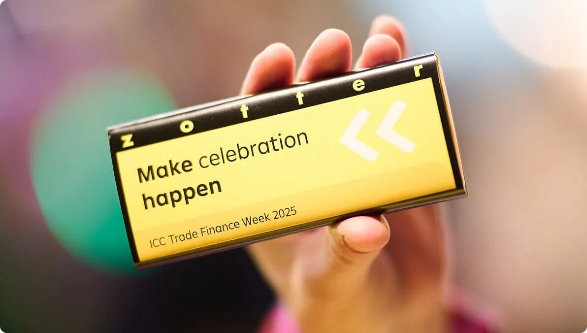 A close-up of a hand holding a Zotter chocolate bar with visible text reading 'Make celebration happen' and 'ICC Trade Finance Week 2025.' The setting features a blurred, colorful background, suggesting an event or festive atmosphere. The packaging is primarily yellow with bold typography and directional graphics.