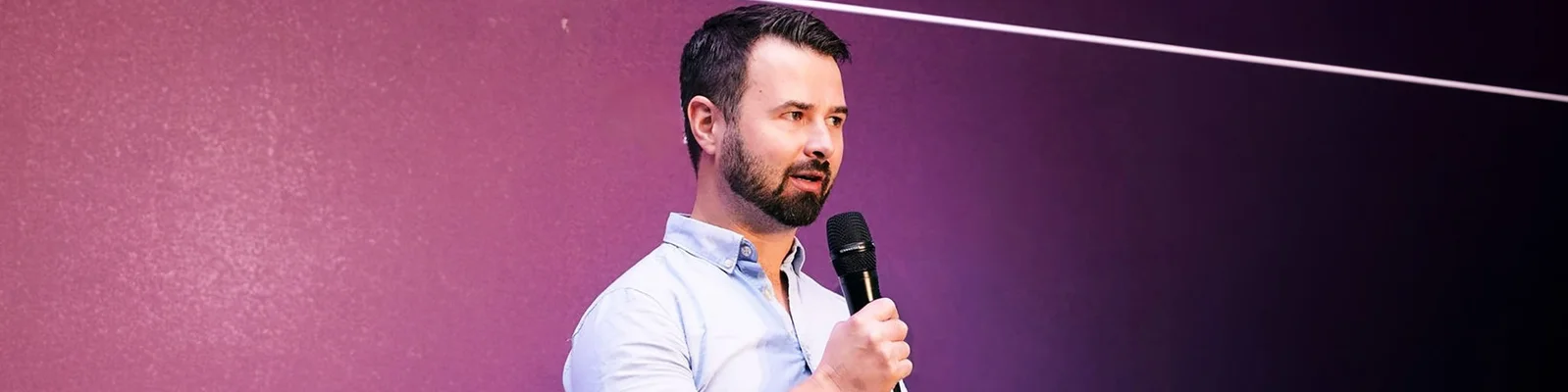 A man with a microphone poses against a vibrant purple wall, ready to engage his audience with his voice.