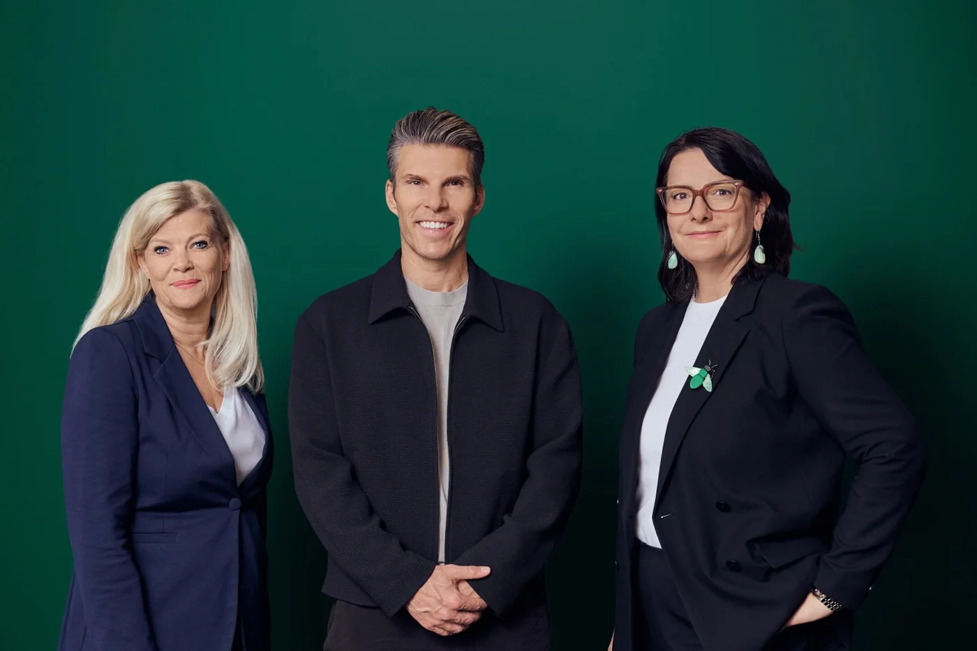 Drei Erwachsene stehen in einem Studiofoto vor einem einfarbig grünen Hintergrund. Sie tragen elegante Businesskleidung, bestehend aus Blazer und legerer Jacke, was auf ein professionelles oder geschäftliches Umfeld hindeutet. Die Person in der Mitte lächelt direkt in die Kamera, während die beiden anderen selbstbewusst an ihrer Seite posieren. Die Gesamtwirkung ist formell und gleichzeitig zugänglich, mit schlichter, klarer Beleuchtung und Komposition.