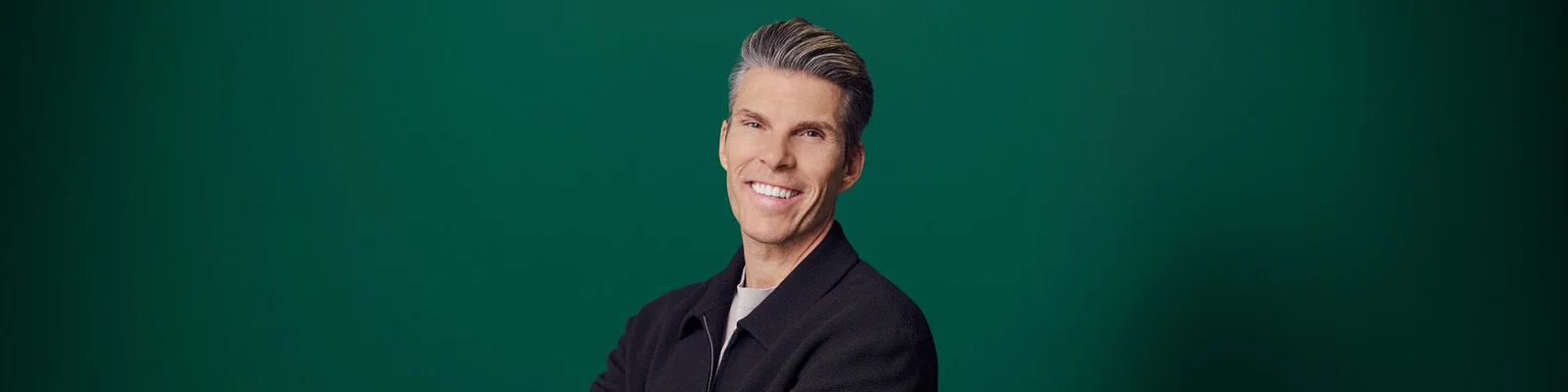 A man with neatly styled gray hair smiles confidently at the camera. He is wearing a dark jacket over a light shirt, giving a polished and professional appearance. The background is a solid green color, creating a clean and modern studio look. The composition focuses on his upper body and friendly expression.