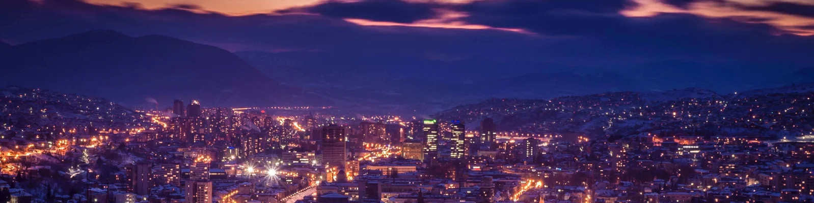 Ein Panoramablick auf eine Stadt, die in der Abenddämmerung von leuchtenden Lichtern erhellt wird. Im Hintergrund erhebt sich eine Bergkulisse unter einem dramatischen, violett-orangenen Himmel. Die Stadtlandschaft präsentiert Gebäude und Straßen, die im Abendlicht erstrahlen und eine zugleich ruhige und lebendige Atmosphäre schaffen.