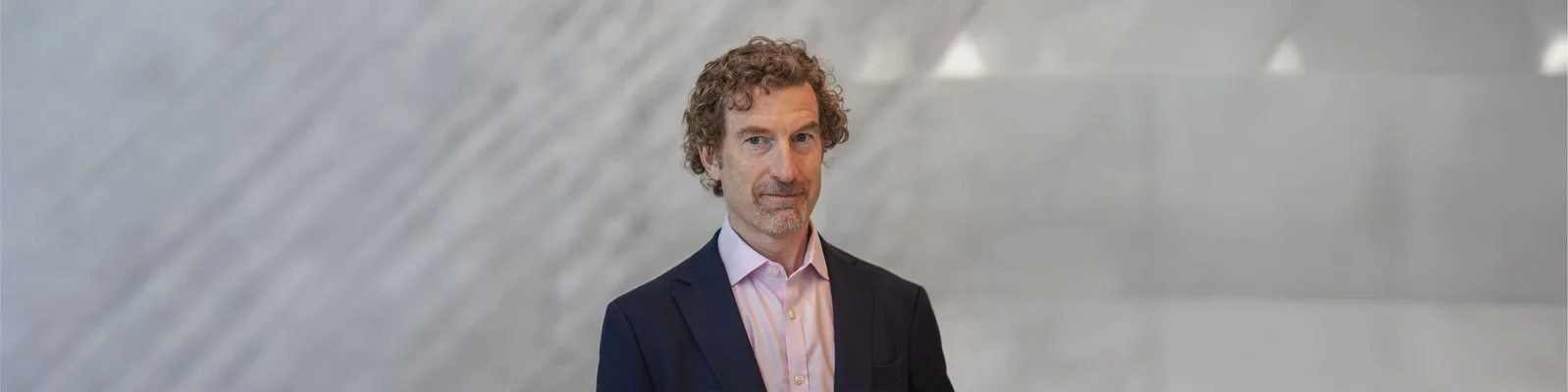 Martin Blum dressed in a dark suit jacket and light pink shirt, showcasing curly hair. The background is a soft, blurred gray pattern.
