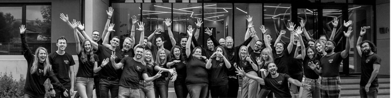 Ein dynamisches Gruppenfoto von Teammitgliedern, die zusammen feiern, mit erhobenen Händen in Freude. Das Bild ist in Schwarzweiß und fängt eine lebhafte und begeisterte Atmosphäre ein.