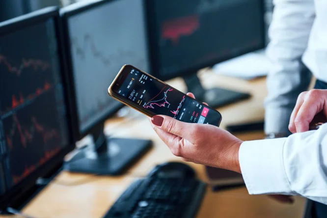 Dieses Bild zeigt eine Person, die ein Smartphone mit Börsendaten in der Hand hält. Im Hintergrund sind auf mehreren Monitoren schwankende Marktdiagramme zu sehen. Es veranschaulicht die Dynamik des Echtzeithandels und der Marktanalyse im Finanzsektor.