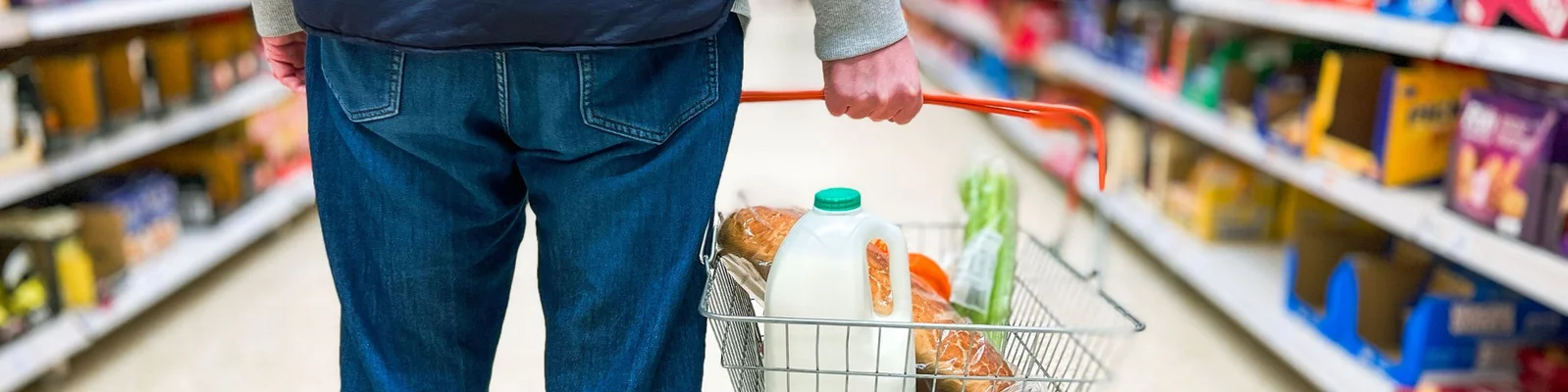 Eine Person, die einen Einkaufswagen mit Lebensmitteln, einschließlich Milch, Brot und Gemüse, hält, während sie durch einen Gang im Supermarkt geht.