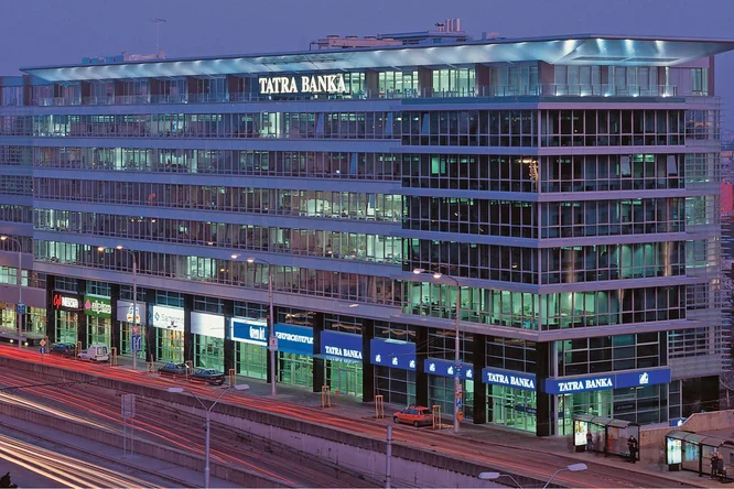 Abendansicht der Tatra Banka Zentrale mit moderner Glasfassade, beleuchtet durch Innenlichter, und Stadtverkehr im Vordergrund.