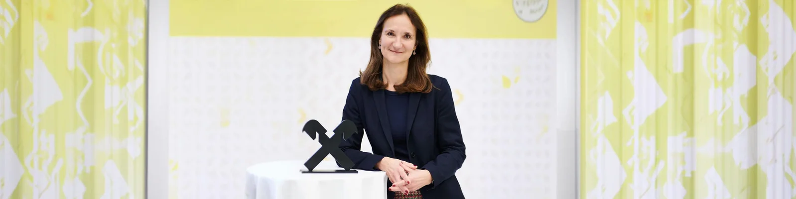 Eine professionell gekleidete Frau steht bei einer Preisverleihung hinter einem kleinen runden Rednerpult. Sie trägt einen dunklen Blazer und stützt die Hände neben einer stilisierten schwarzen Trophäe auf das Pult. Im Hintergrund ist eine leuchtend gelb-weiße Grafikwand mit dezenten Mustern und Textelementen zu sehen. Die Szene wirkt wie eine formelle Auszeichnungsveranstaltung eines Unternehmens oder einer Branche.