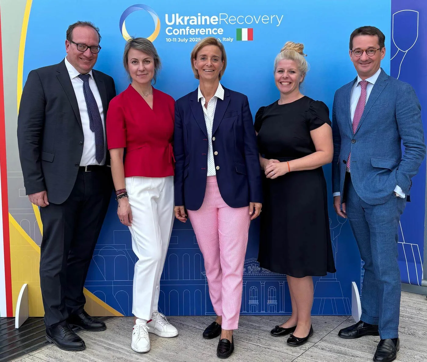 A group of five individuals stands in front of a blue backdrop featuring the 'Ukraine Recovery Conference' text, dated '10-11 July 2023' and located in Italy. The backdrop includes a circular graphic and the Italian flag. The individuals are dressed in formal and semi-formal attire, with a mix of vibrant and neutral tones.