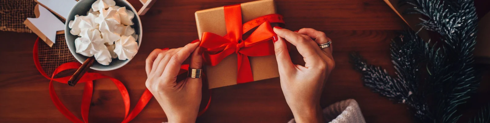 Hände binden ein rotes Band um eine braune Geschenkbox, umgeben von Weihnachtsdekoration, Kerzen und einer Schale mit Baisers.
